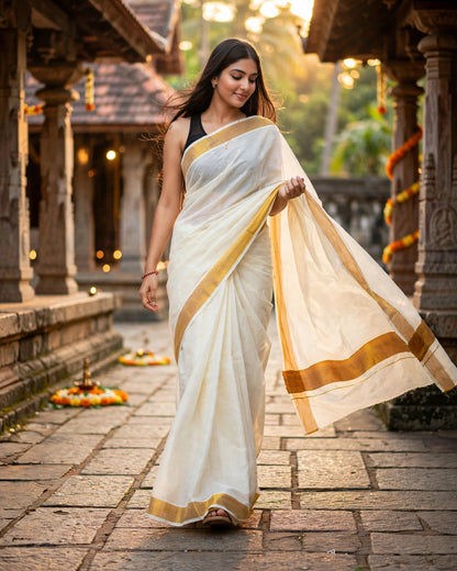 A woman wearing a white saree with gold borders walks through a stone corridor at sunset. The saree has a black blouse underneath, and the background features warm lighting and traditional architectural details.